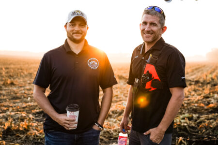 Luft and Sons standing in a harvested field during sunset, one holding a coffee cup and the other a water bottle, both wearing casual work attire with company logos.