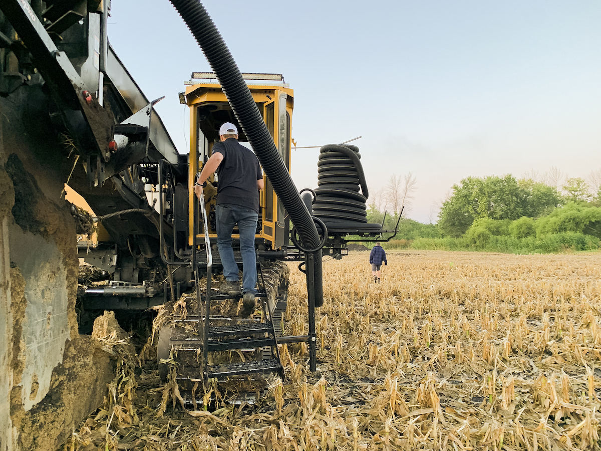 An agricultural scene with a Luft and Sons worker climbing the steps into a yellow Wolfe trenching machine, with large coils of black flexible piping loaded on the back. In the background, another person can be observed walking away across the harvested field.