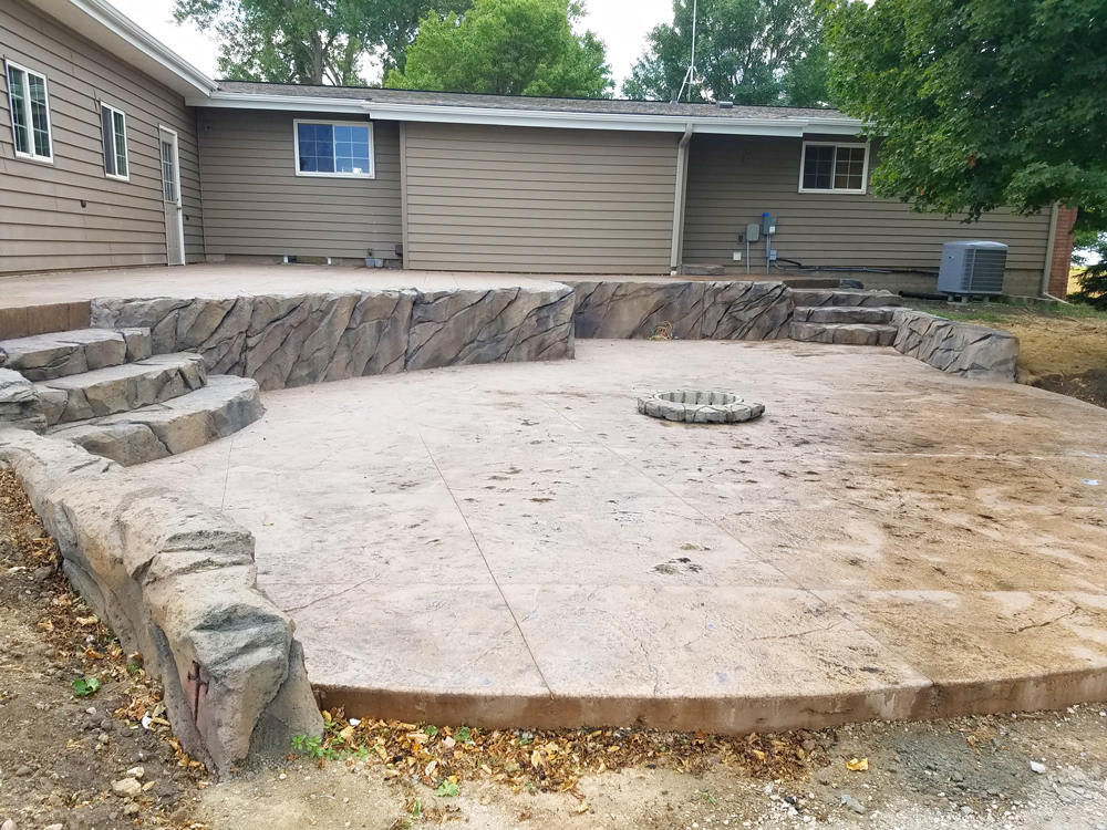 A spacious outdoor patio made with stamped concrete and bordered by a natural stone retaining wall, with integrated steps leading to a residential backyard. In the center is a small circular fire pit.