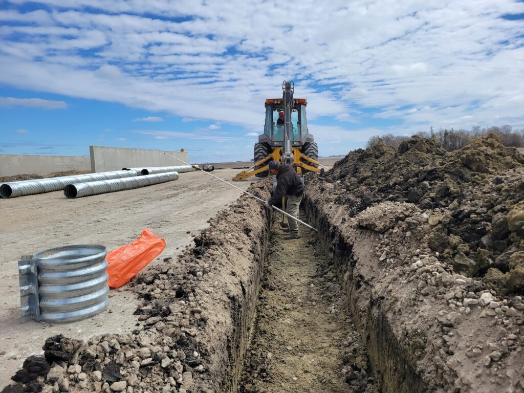 A Luft and Sons construction worker in a black jacket uses a measuring tape in a long, narrow trench with corrugated pipes nearby, while a yellow backhoe stands ready, under a partly cloudy sky.
