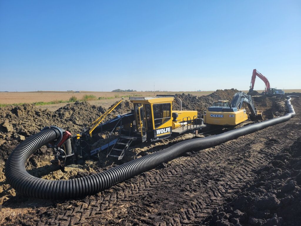 Agricultural landscape with Luft and Sons yellow trenching machine laying large black flexible piping in the soil, with a red excavator in the background under a clear blue sky.