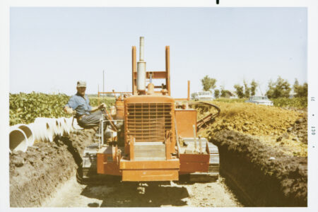 Vintage photo of a Luft and Sons worker operating an orange trencher creating a deep trench in a field, with white corrugated pipes laid out to the side under a clear sky.