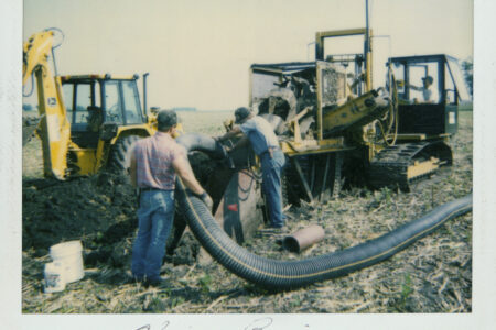 Luft and Sons workers in casual clothing are seen around a trenching machine, handling large black flexible pipes on a farm, with a yellow digger in the background.